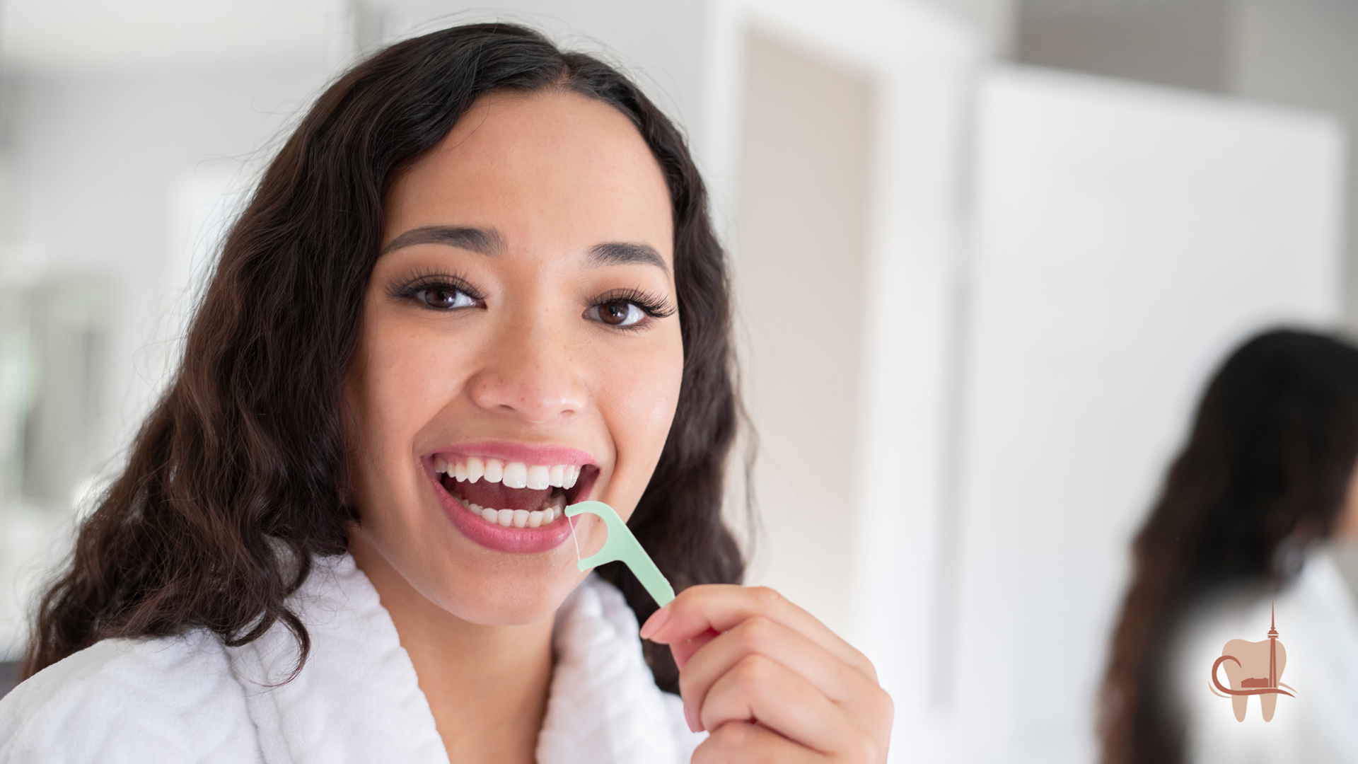Woman flossing teeth in bathroom, wearing a robe, smiling.
