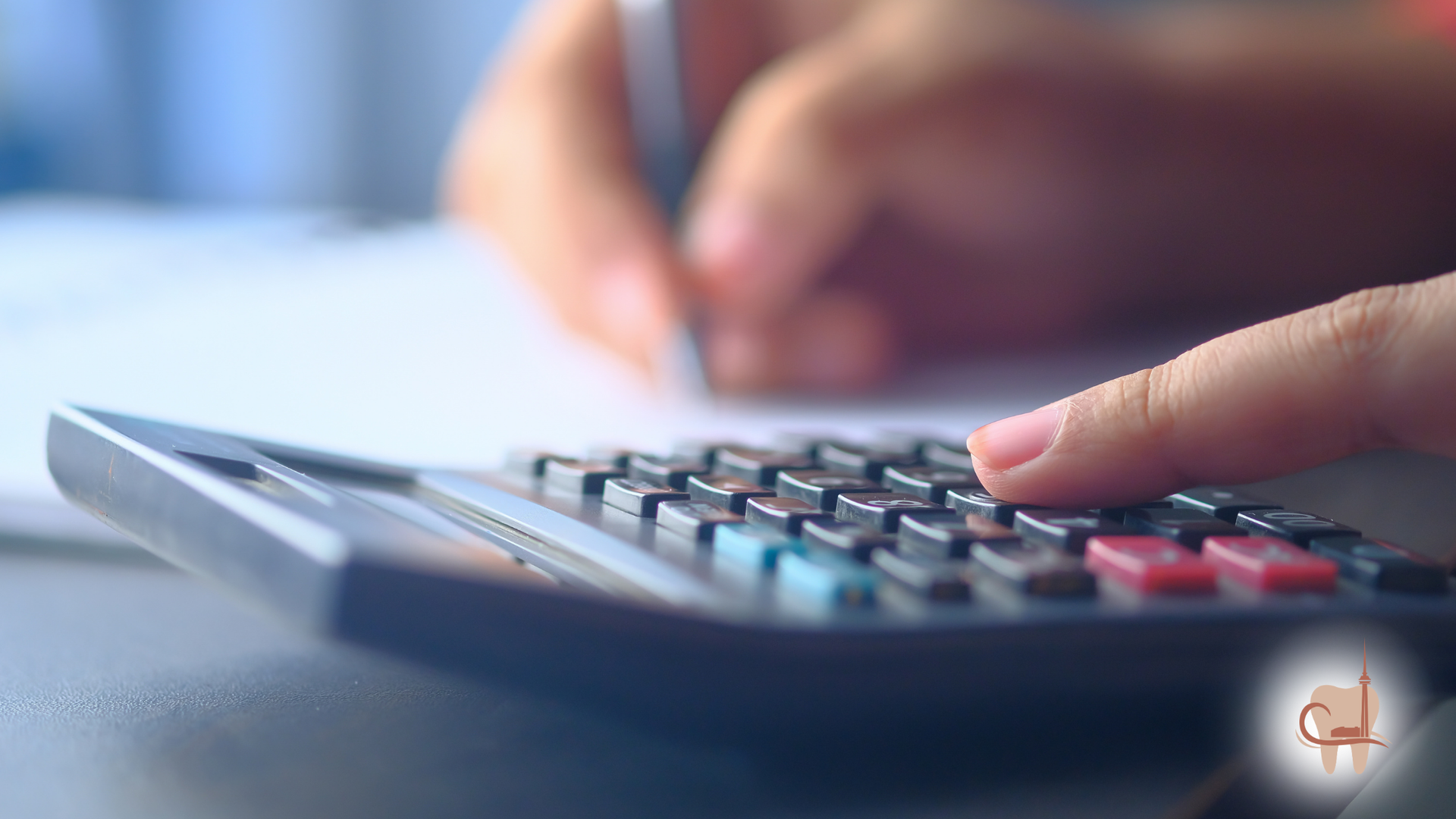 Person's hand using a calculator; another hand writes on paper, blurred background.