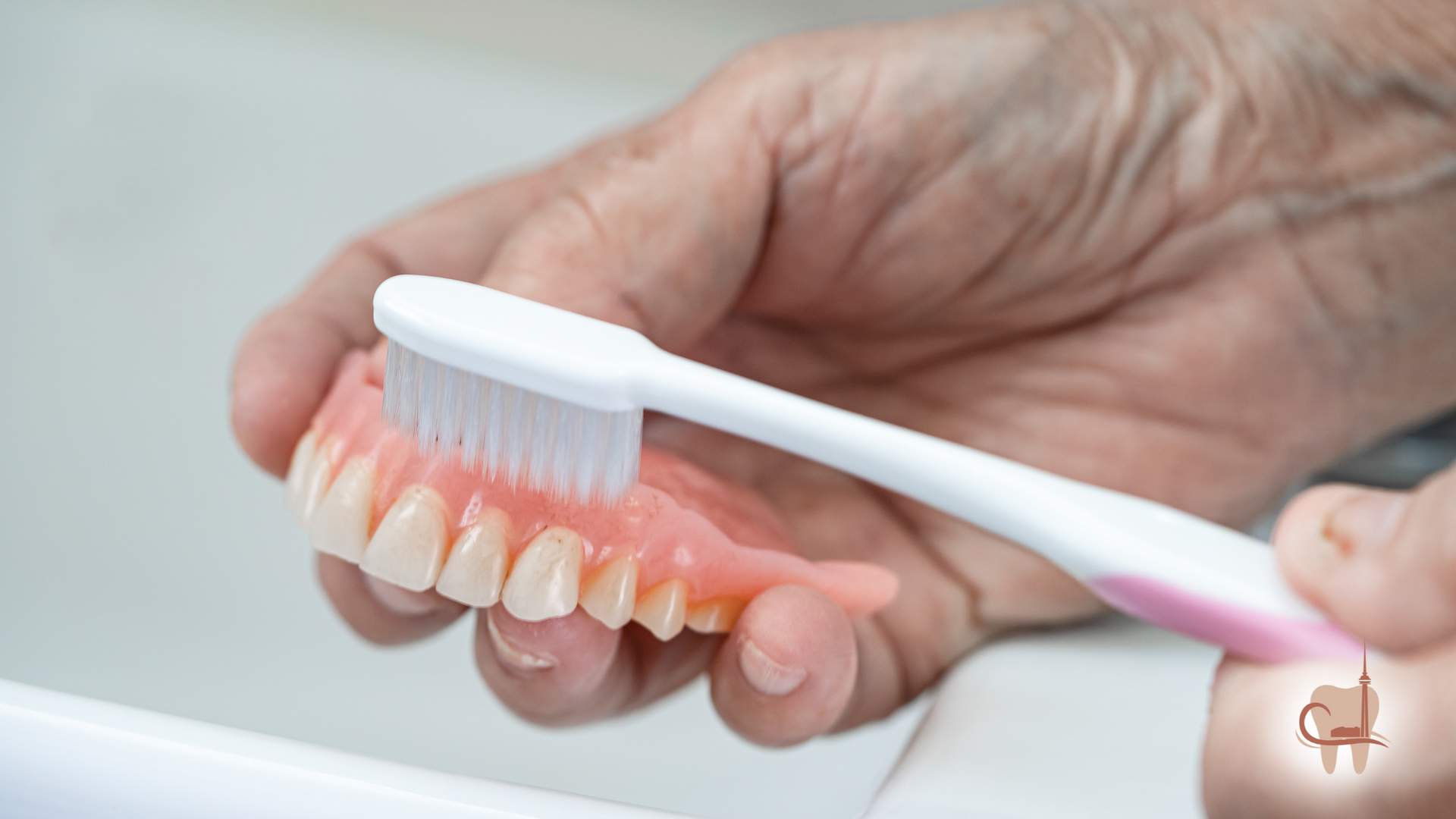 Person cleaning dentures with a toothbrush over a sink; hands visible.