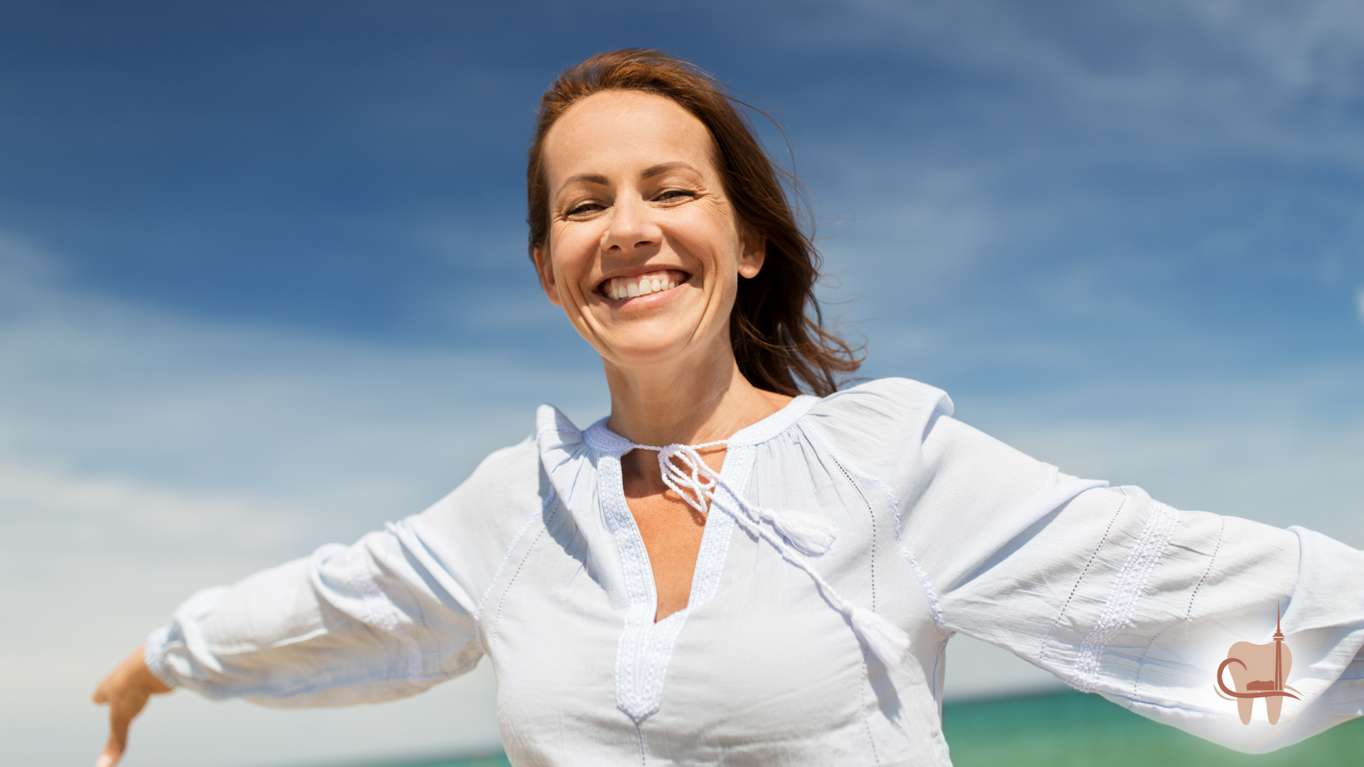 Woman with arms outstretched smiling broadly against a blue sky and ocean.