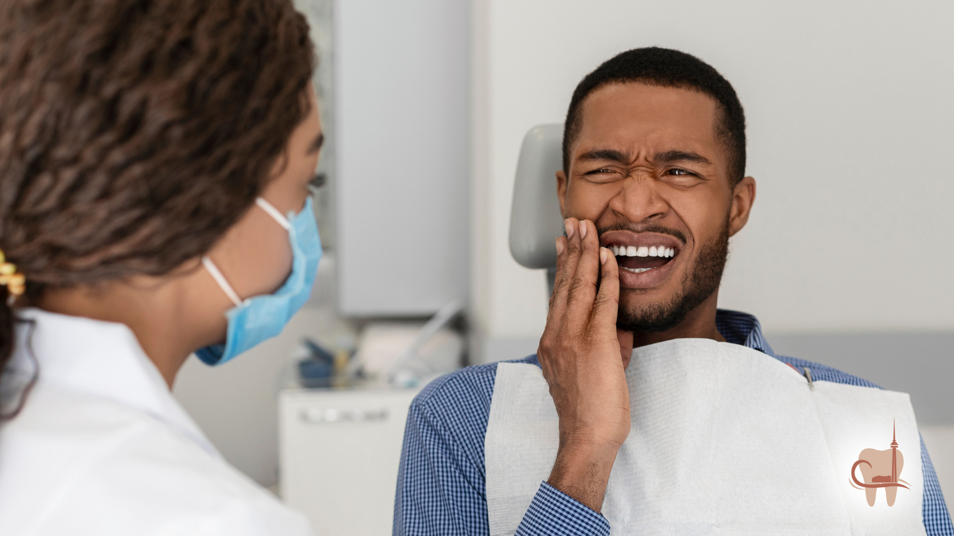 A dentist examining a Black man with a painful expression, holding his jaw in a dental office.