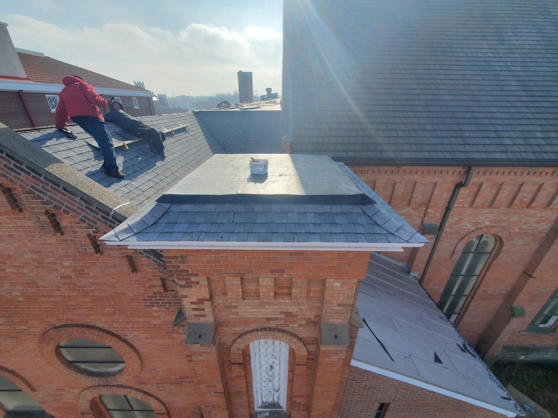 Person on a roof with red jacket, working on shingles, sunny day. Building exterior.