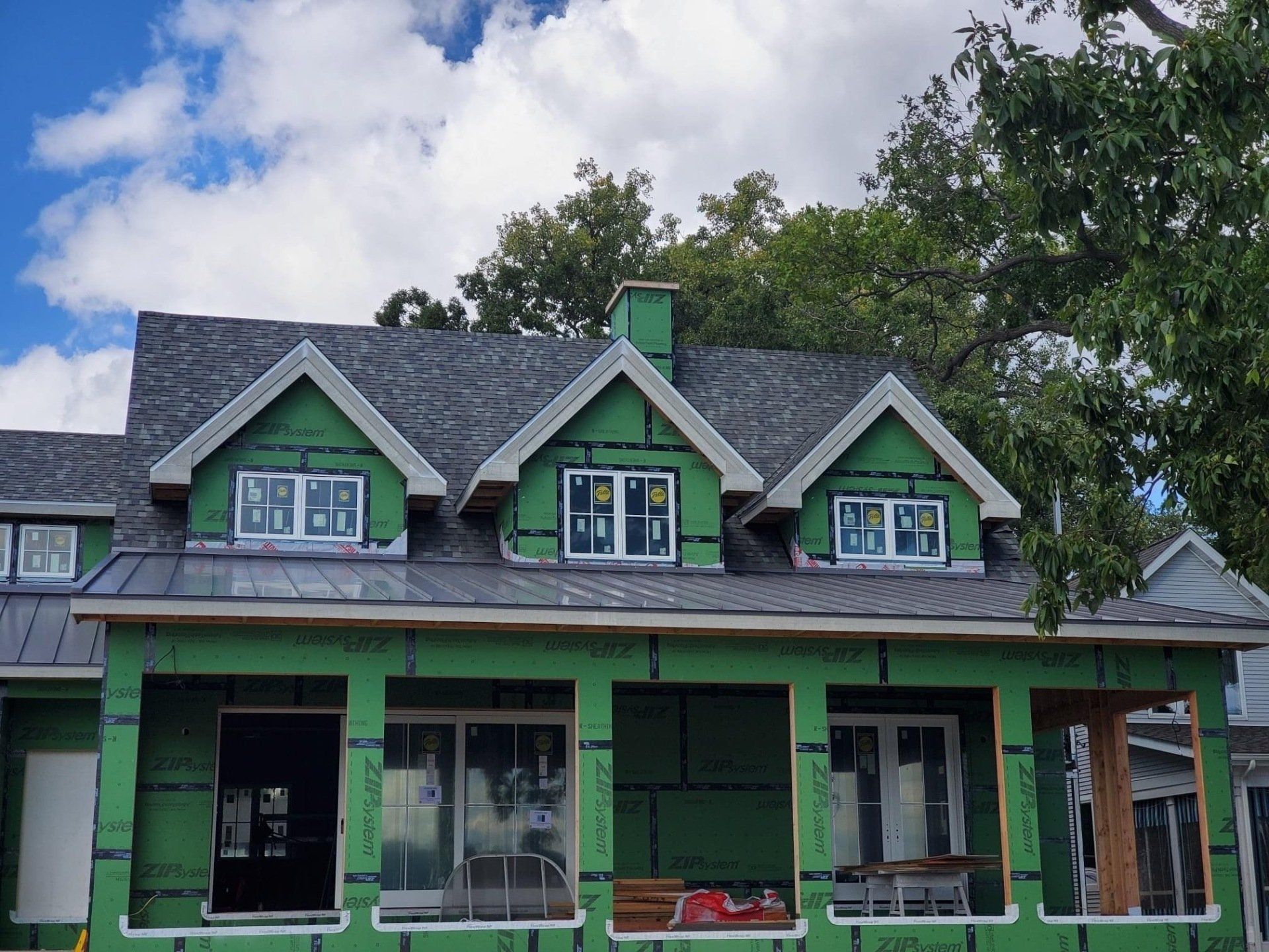 House under construction with green wrap, gray roof, and dormer windows against a cloudy sky.