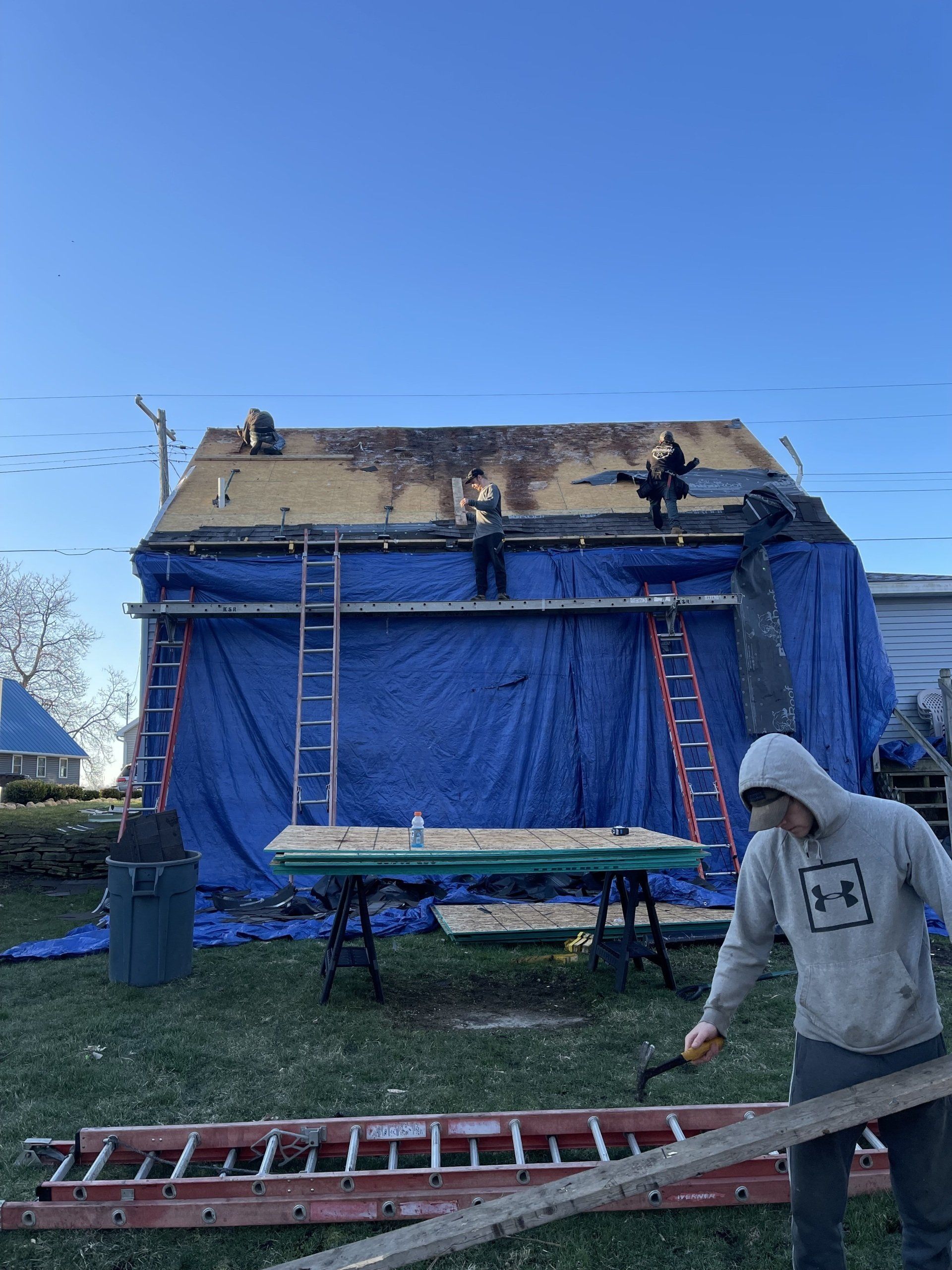 Roofing crew working on a house covered in blue tarp. One man holds a tool, others work on the roof.