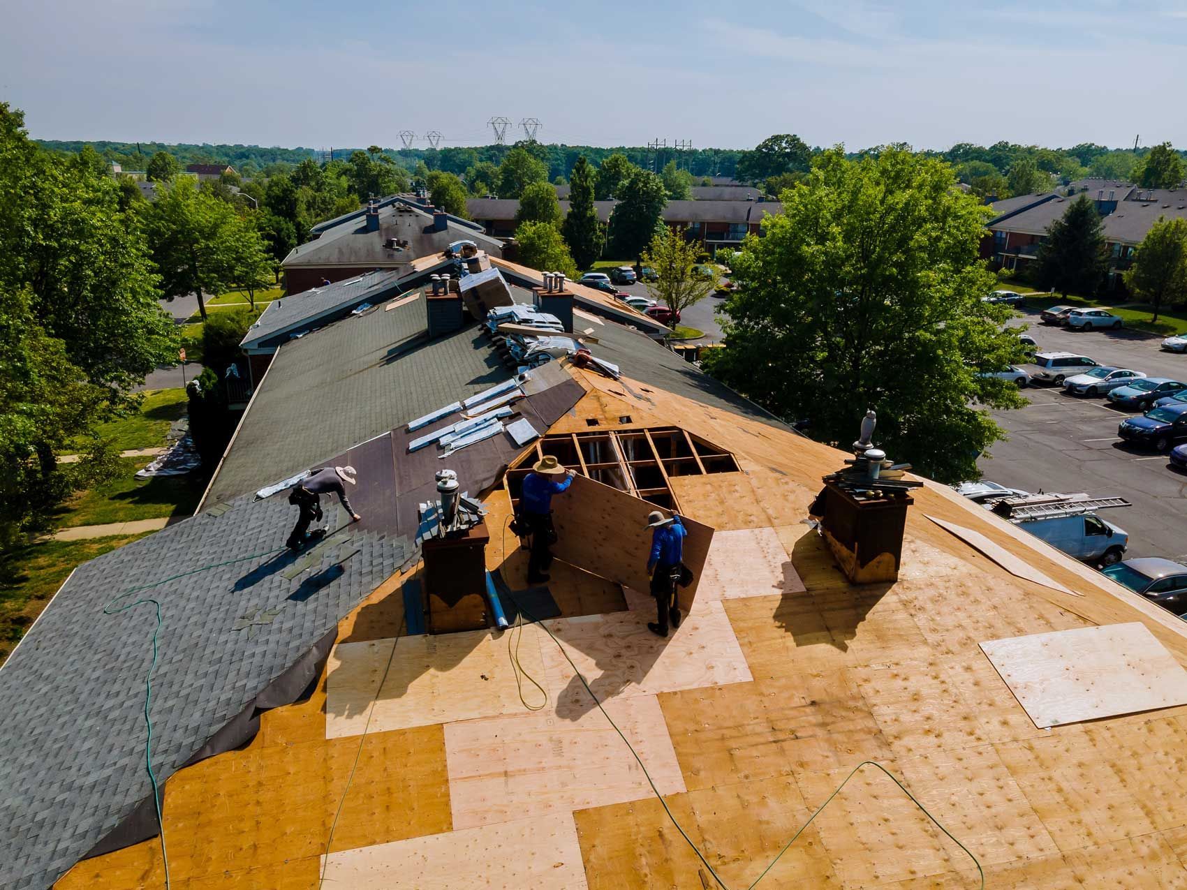 Roofers replacing a shingled roof on a residential building with surrounding trees and parked cars.
