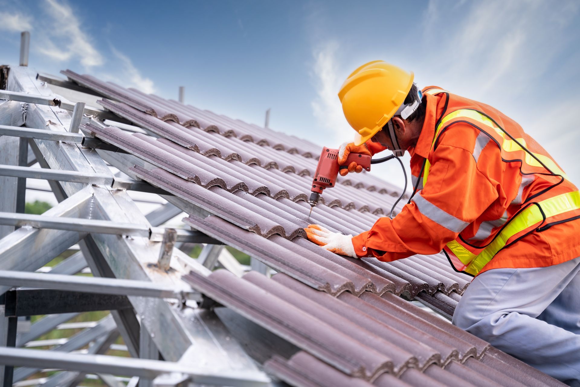 Male roofing contractor repairing tiled roof under sunset sky in safety gear using a power tool.