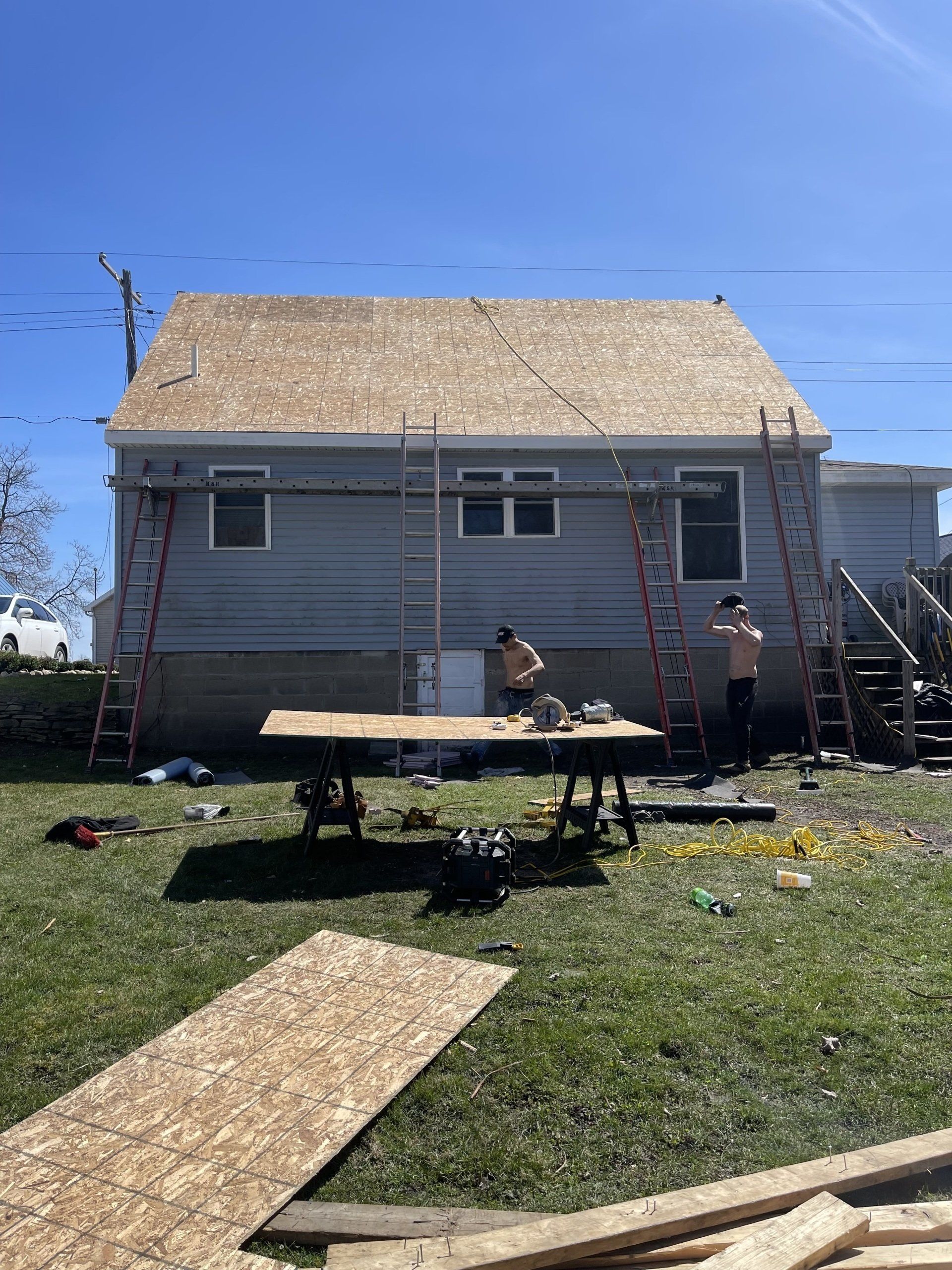 Workers on a roof, installing shingles on a blue-sided house. Ladders and tools are on the ground.