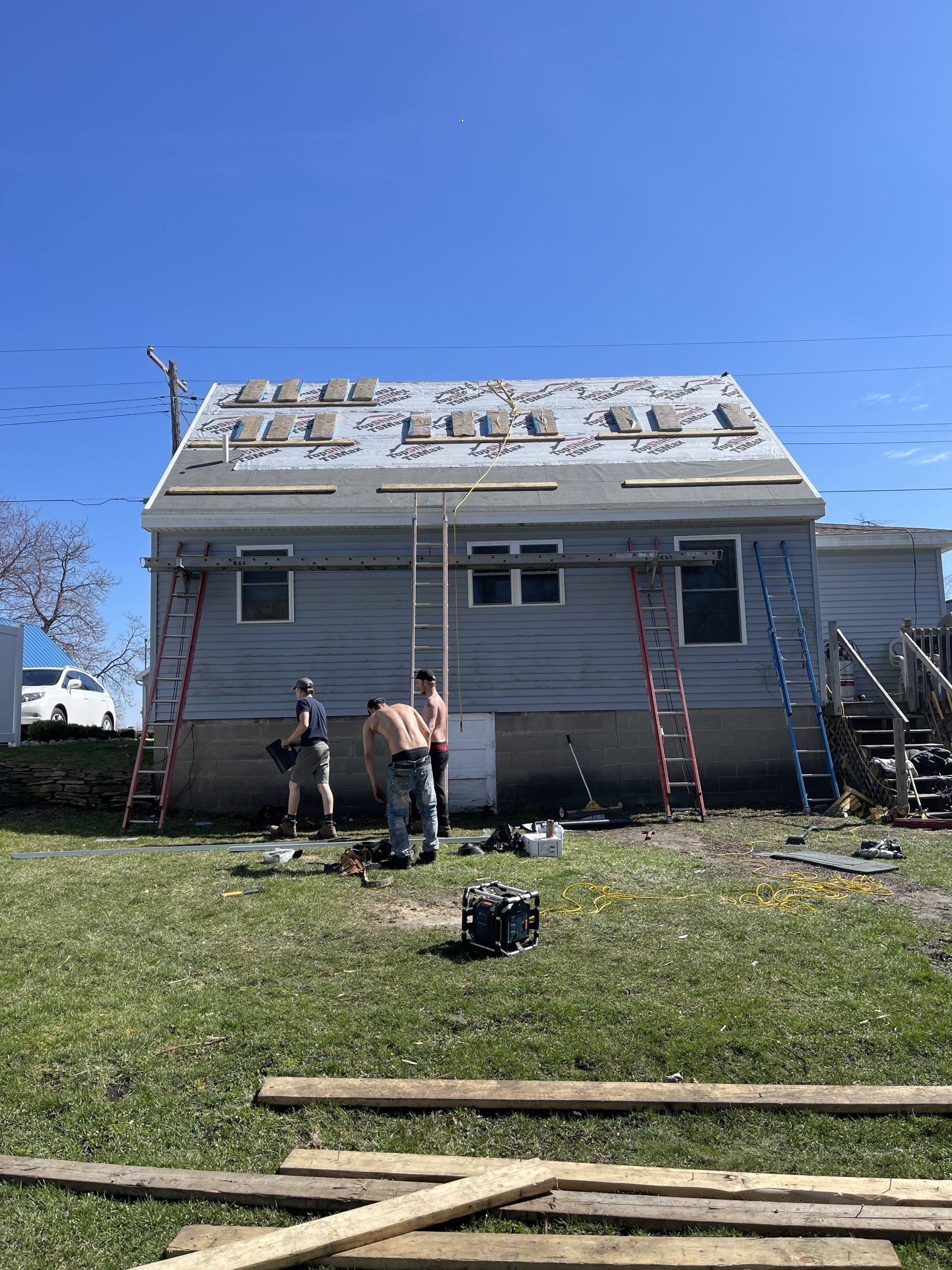 Three people on a lawn working on a house roof on a sunny day. Ladders lean on the house.