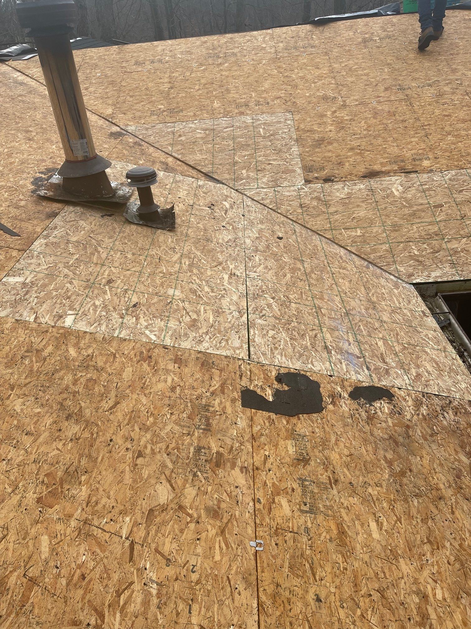 OSB roof sheathing with chimney and vent pipe, viewed from above, with a person in the distance.