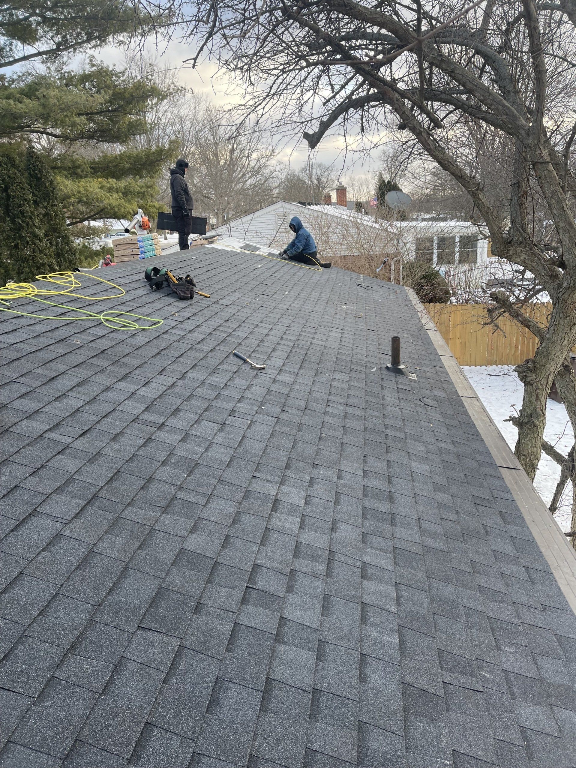 Two people working on a gray shingle roof in a snowy setting.