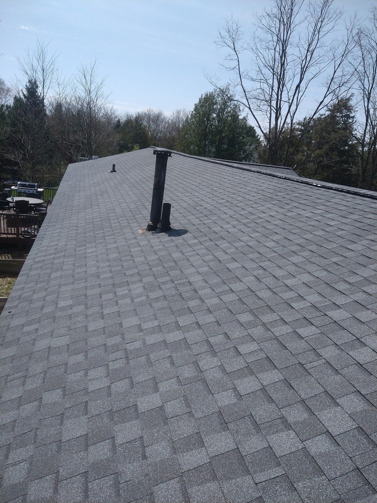 Gray shingle roof with black pipes against a bright sky, trees in the background.
