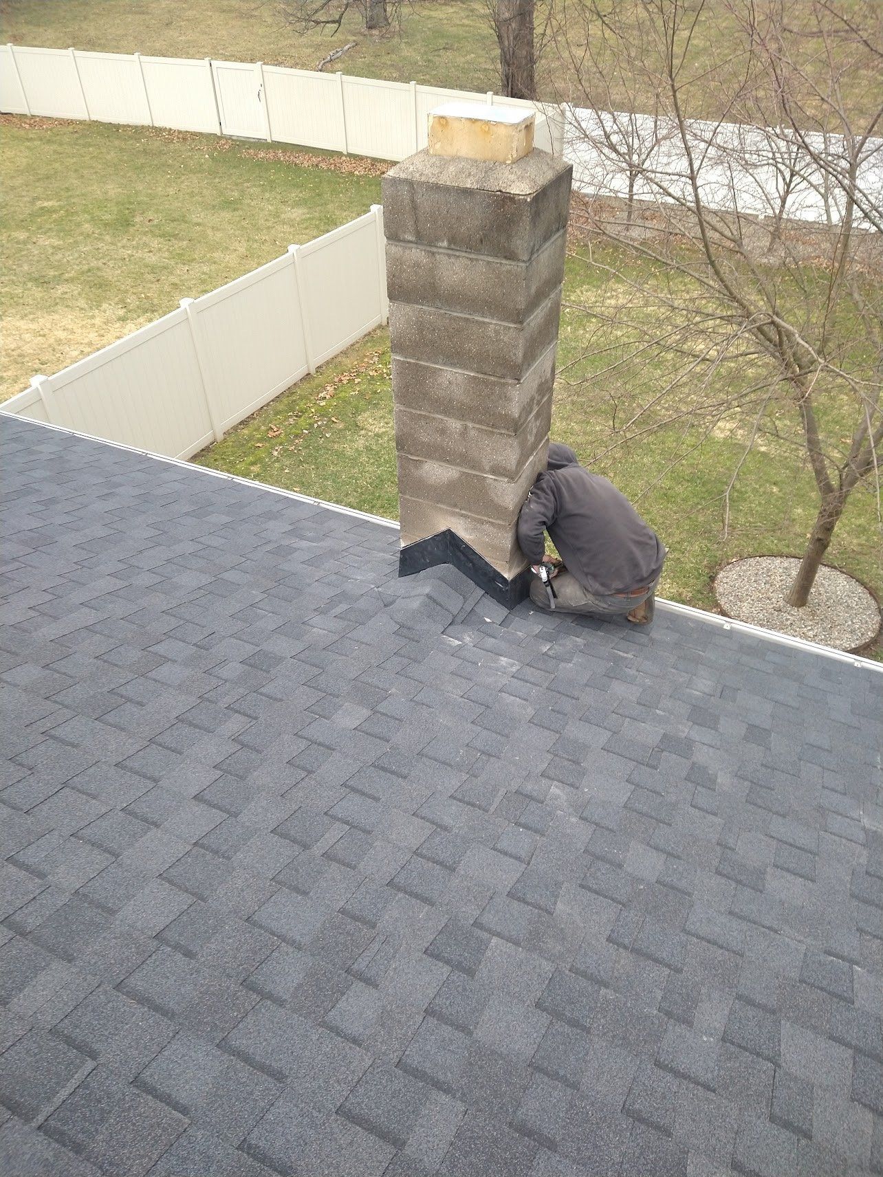 Person repairing flashing around a chimney on a gray asphalt shingle roof.