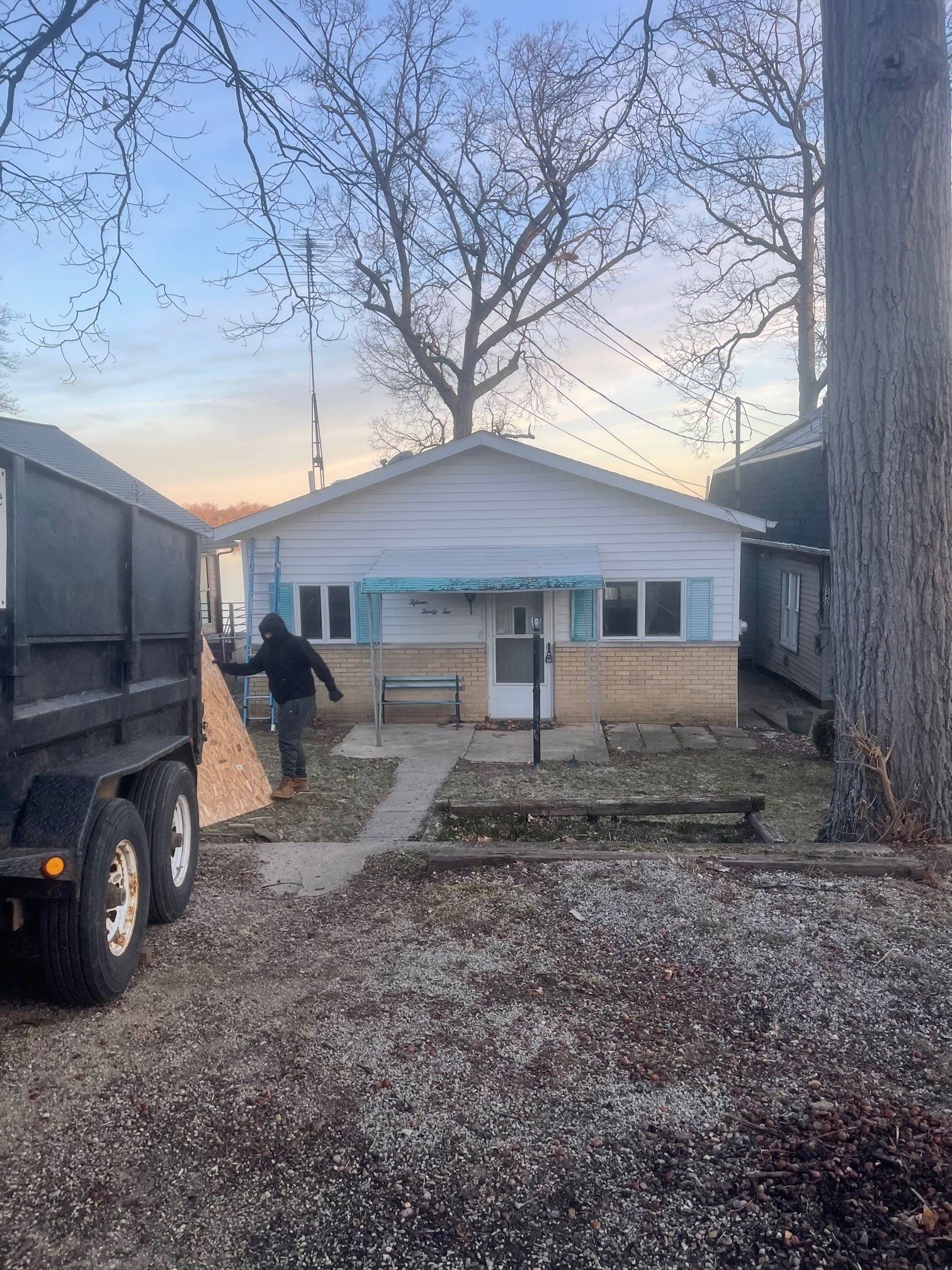 Small white house with blue trim, person near open trailer. Trees and overcast sky.