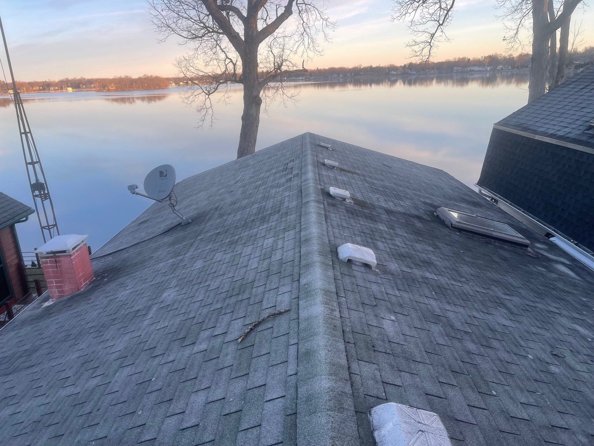 Gray shingled roof overlooking a calm body of water at dusk, with trees in the background.