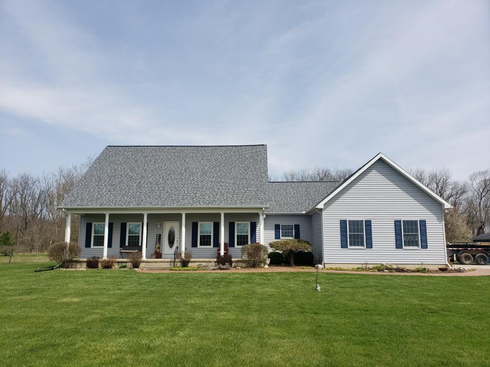 A light gray ranch-style house with blue shutters, a porch, and a green lawn under a blue sky.