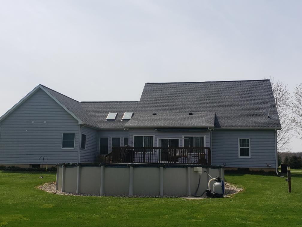 Backyard with above-ground pool and gray house with deck and dark roof under a bright sky.