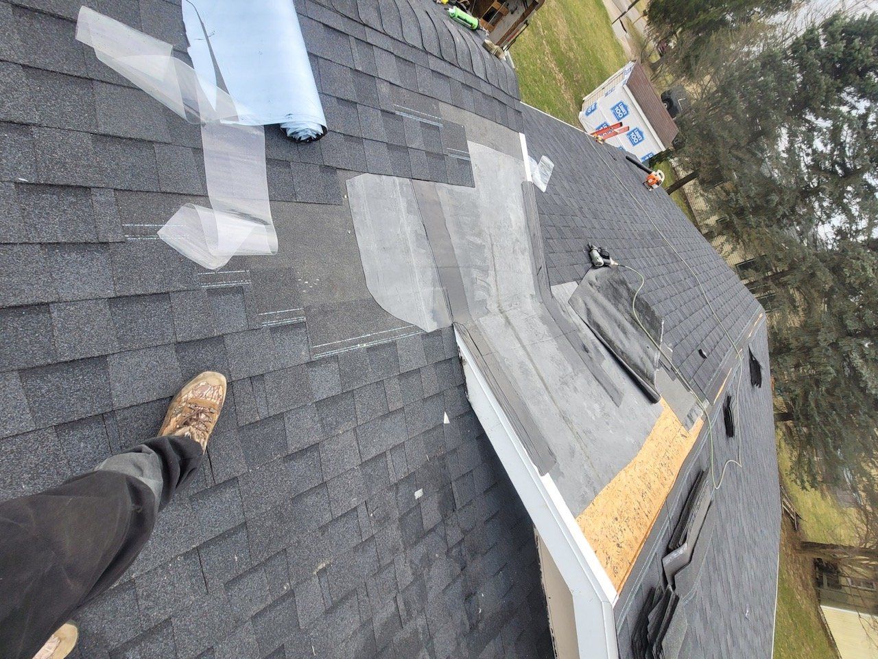 Person standing on a dark asphalt shingle roof, with repair materials visible.