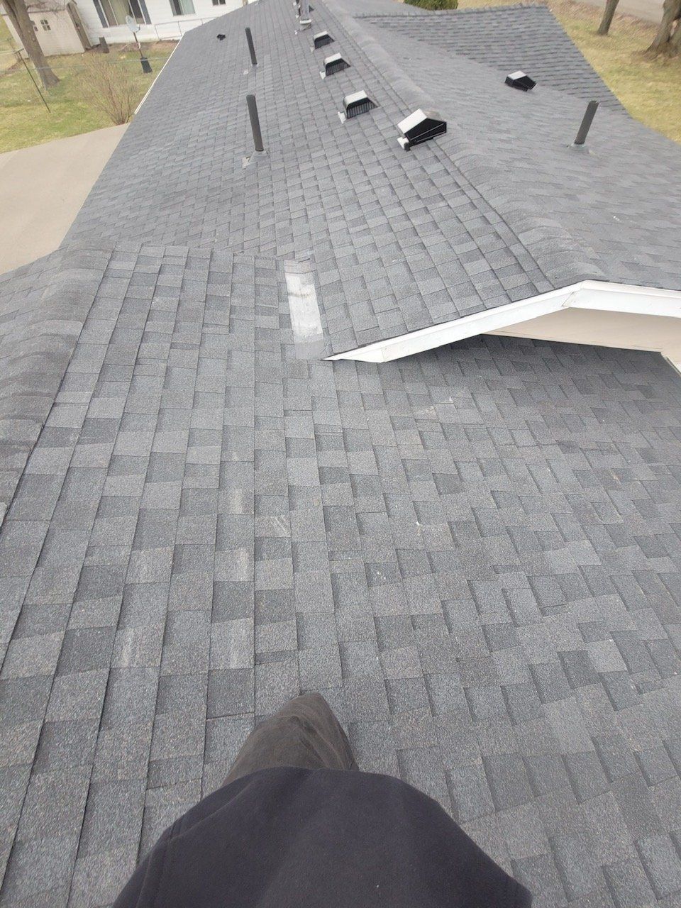 Person standing on a dark asphalt shingle roof. Long roof with multiple vents and a white-trimmed gable.