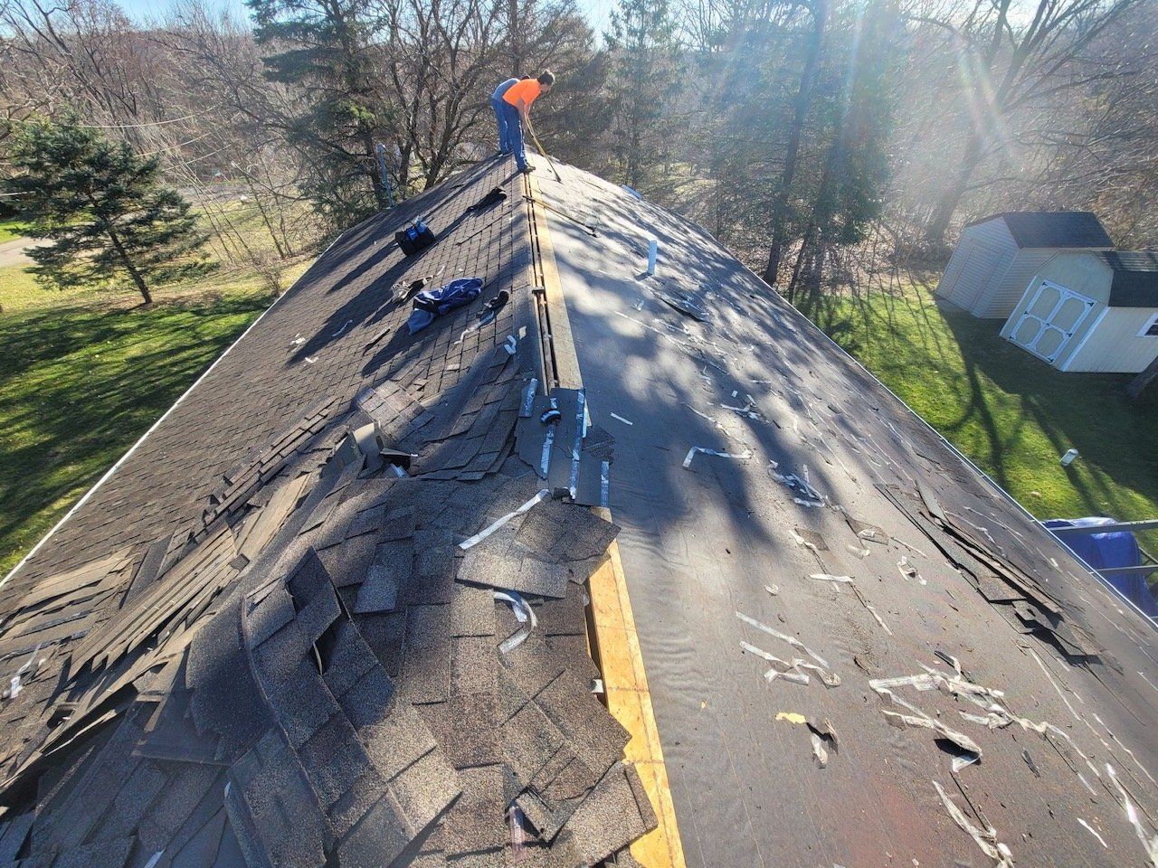 Roofer standing on a roof with damaged shingles. Trees and a house in background, sunny day.
