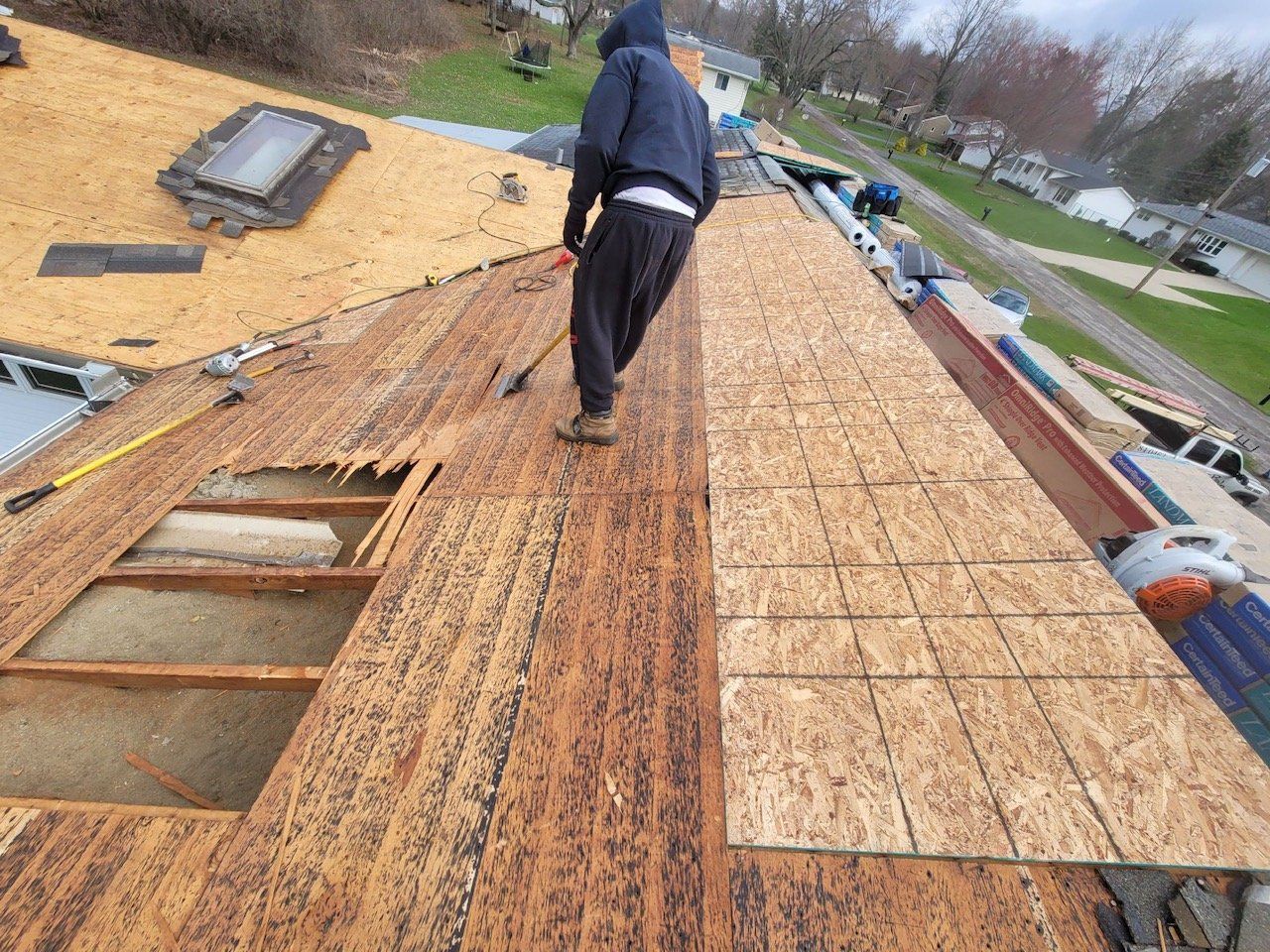 Roofer on a partially torn-off roof, replacing damaged wood panels. Overcast day.