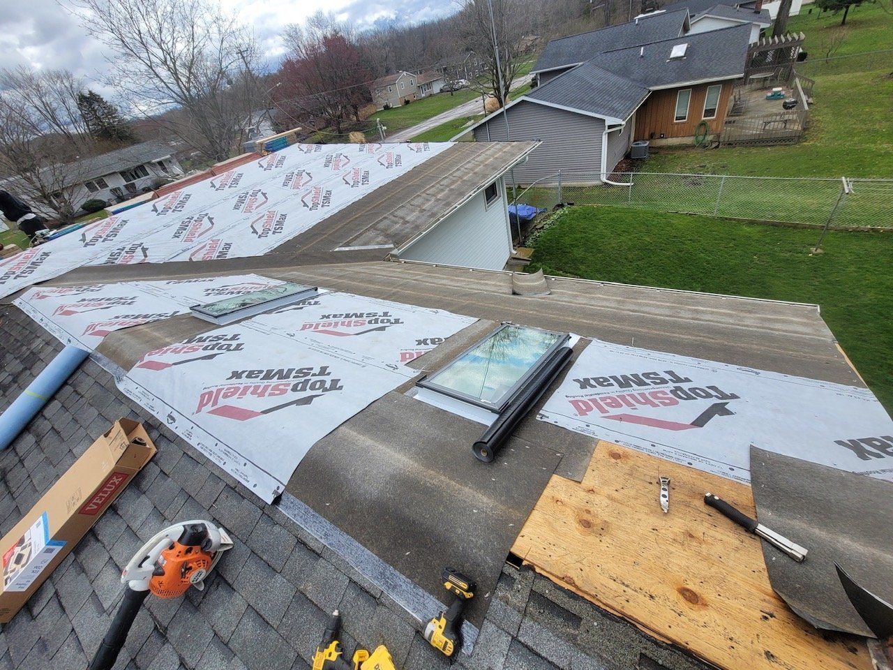Roof partially covered with protective underlayment, skylight visible, tools and supplies scattered.