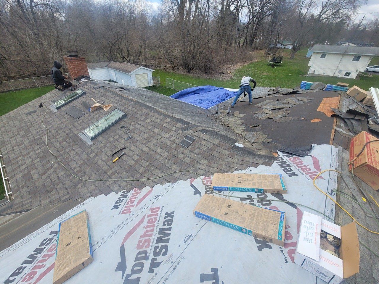Roofers working on a house roof. Blue tarp, materials, and a chimney are visible on the roof.