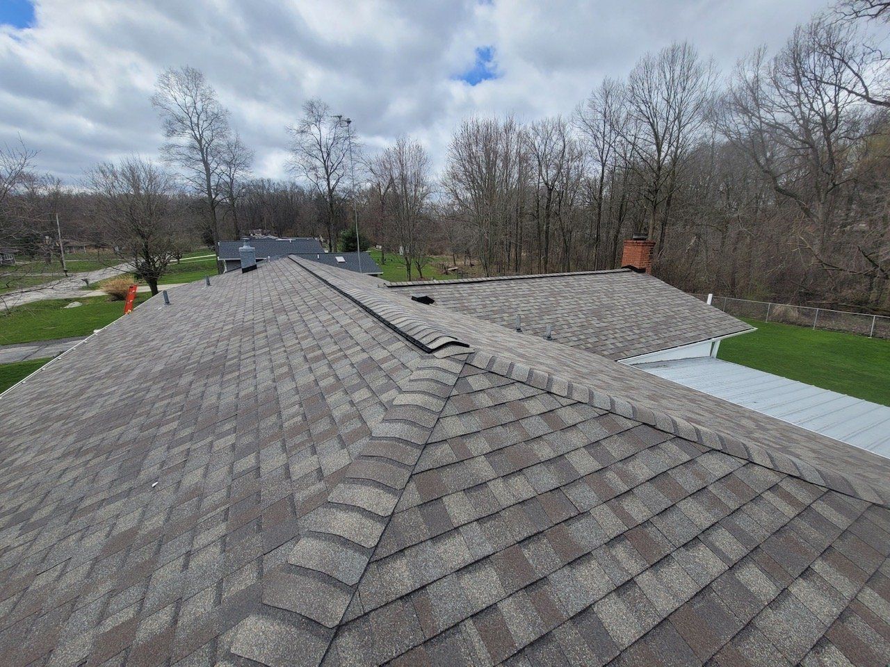 View of a multi-sectioned brown shingled roof with trees and cloudy sky in the background.