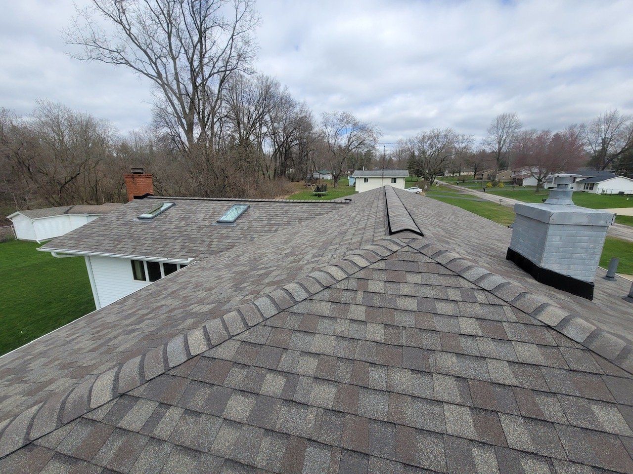 A roof with gray shingles, two skylights, a chimney, and a treeline in the background on a cloudy day.