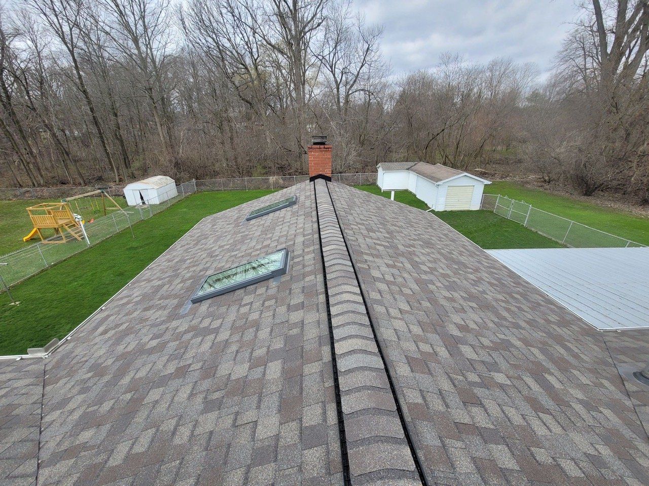 View from a roof with brown shingles, chimney, skylights, and backyard with grass and trees.