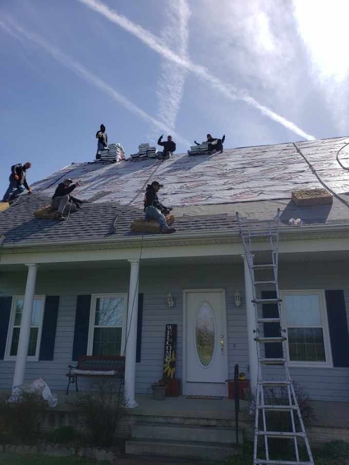 Roofers working on a house roof on a sunny day. Blue sky, several workers visible, and a ladder.
