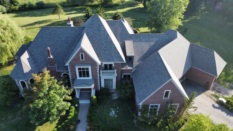 Aerial view of a large brick house with a complex roof and attached garage, surrounded by greenery.