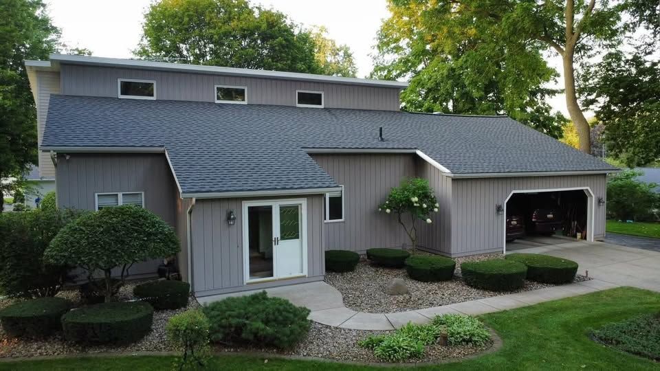 Gray house with dark roof and garage; landscaped yard with shrubs.