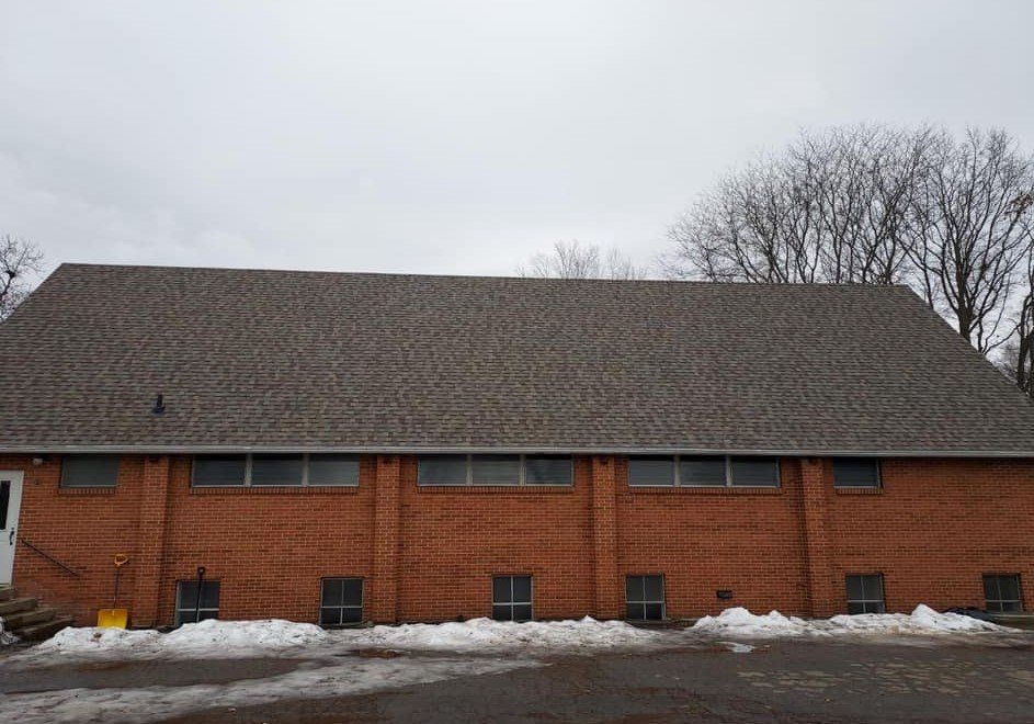 Brick building with a brown shingled roof under a cloudy sky. Small windows and snow in the foreground.