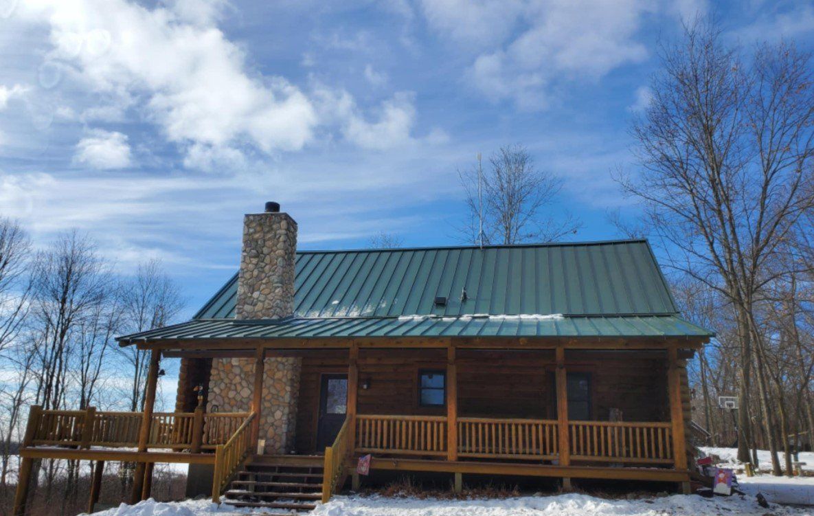 Log cabin with green metal roof, stone chimney, and wooden porch, set in a snowy landscape under a cloudy blue sky.