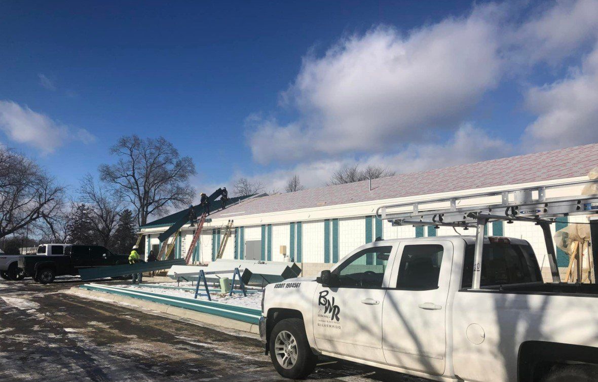 Workers on a roof, installing new shingles. White truck in the foreground, snow on the ground, blue sky.