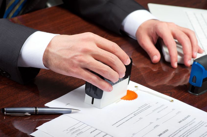 Hands stamping a document on a desk beside a pen and blue stamp tool