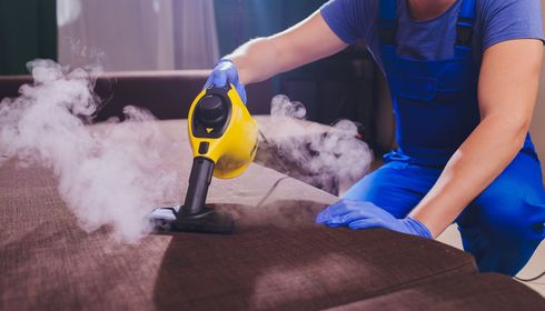 Person in blue overalls steam-cleaning a brown couch with a yellow steam cleaner. Smoke is visible.