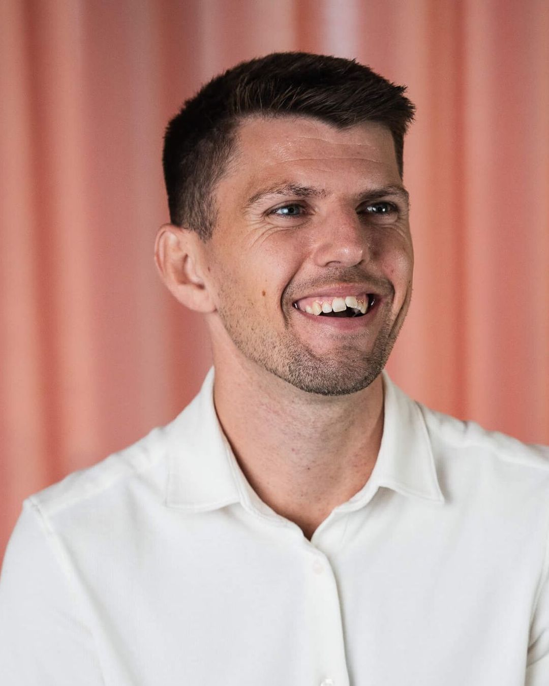 A man in a white shirt is smiling in front of a pink curtain.