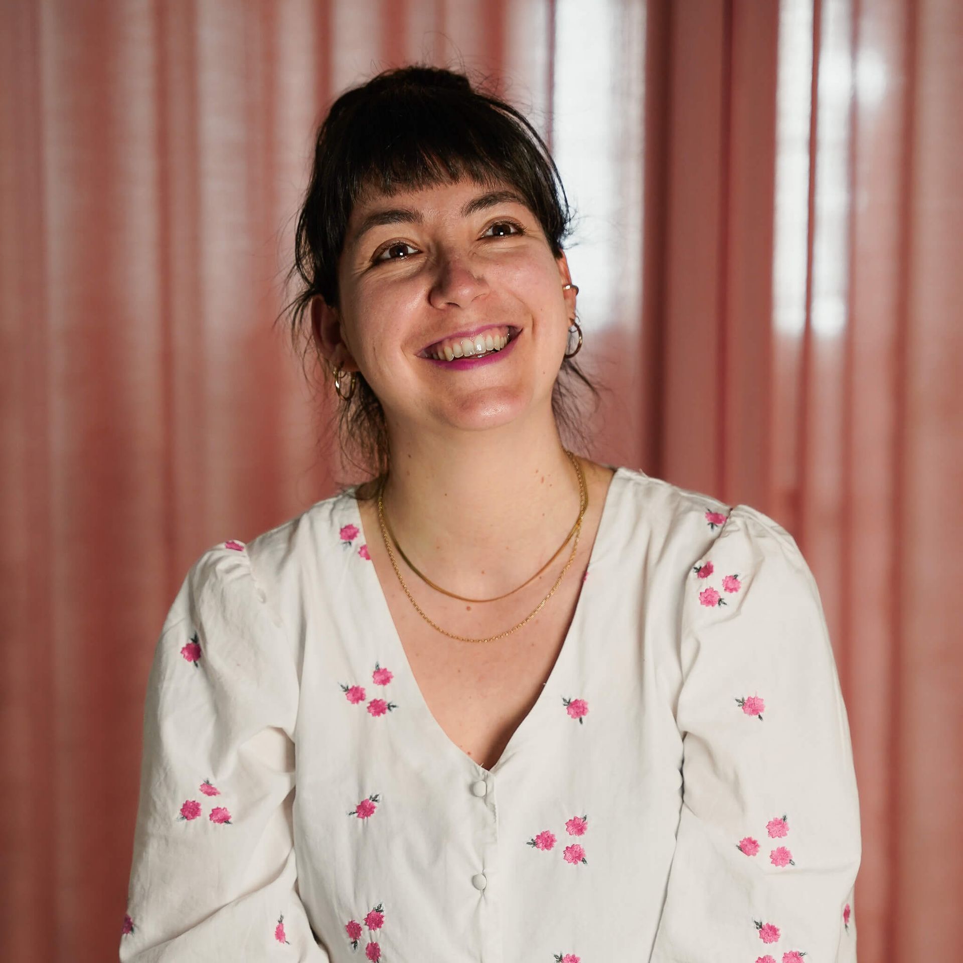A woman in a pink jacket is smiling in front of a pink curtain.