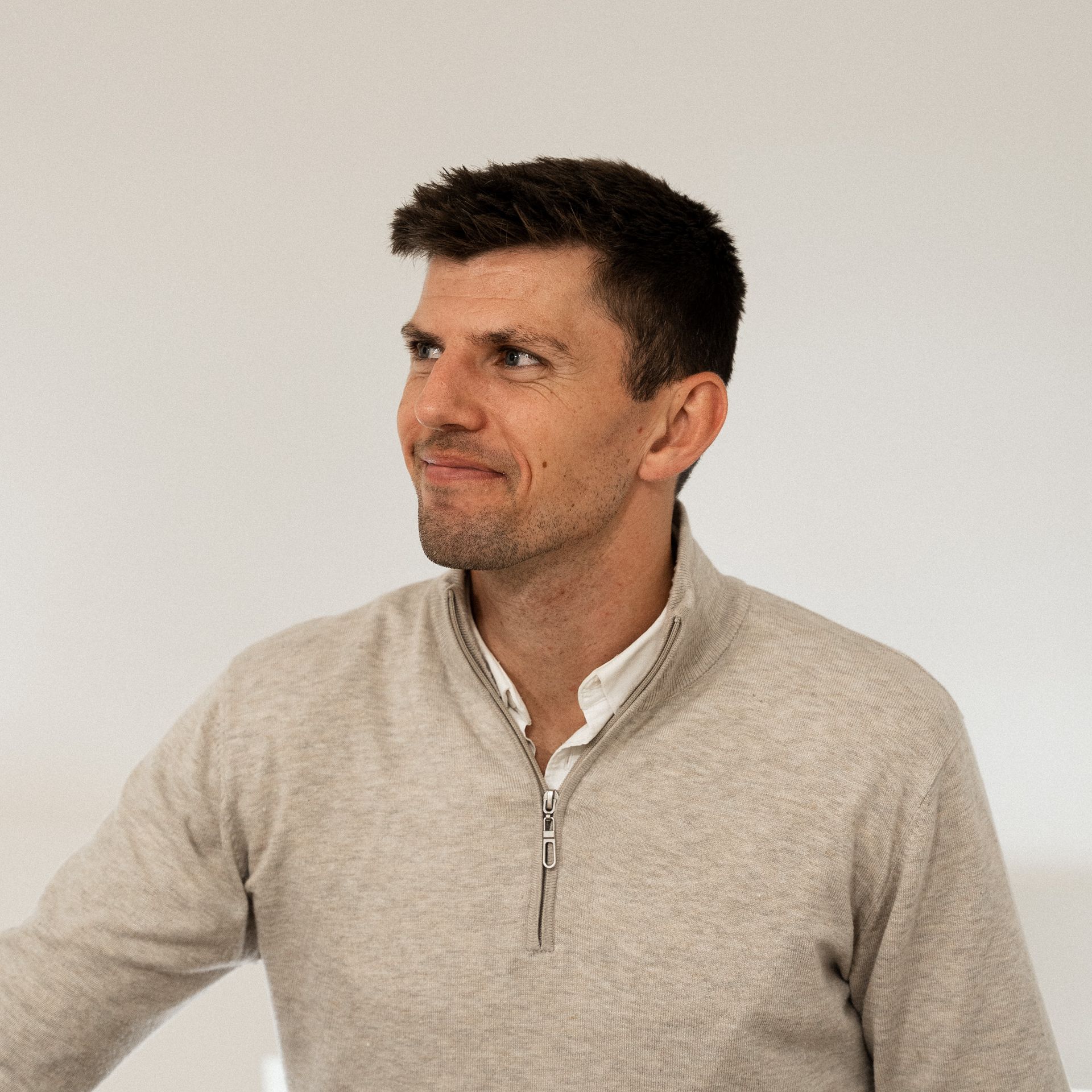 A man wearing a sweater and a white shirt is standing in front of a white wall.