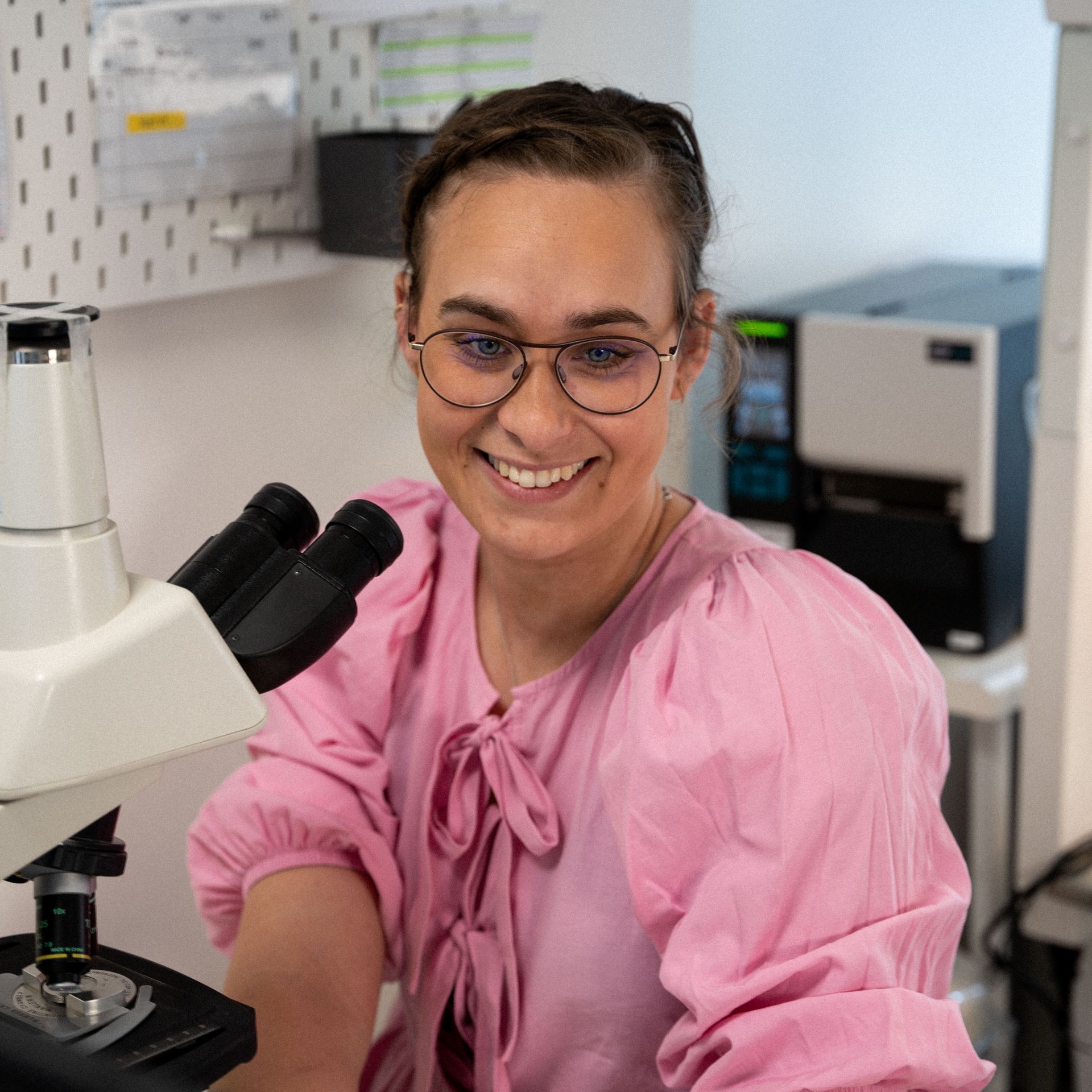 A woman in a pink shirt is smiling while looking through a microscope.