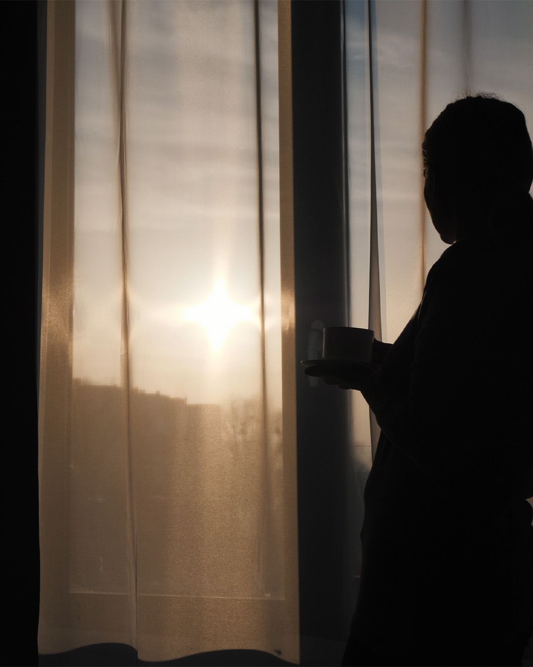 A silhouette of a person standing in front of a window with the sun shining through the curtains