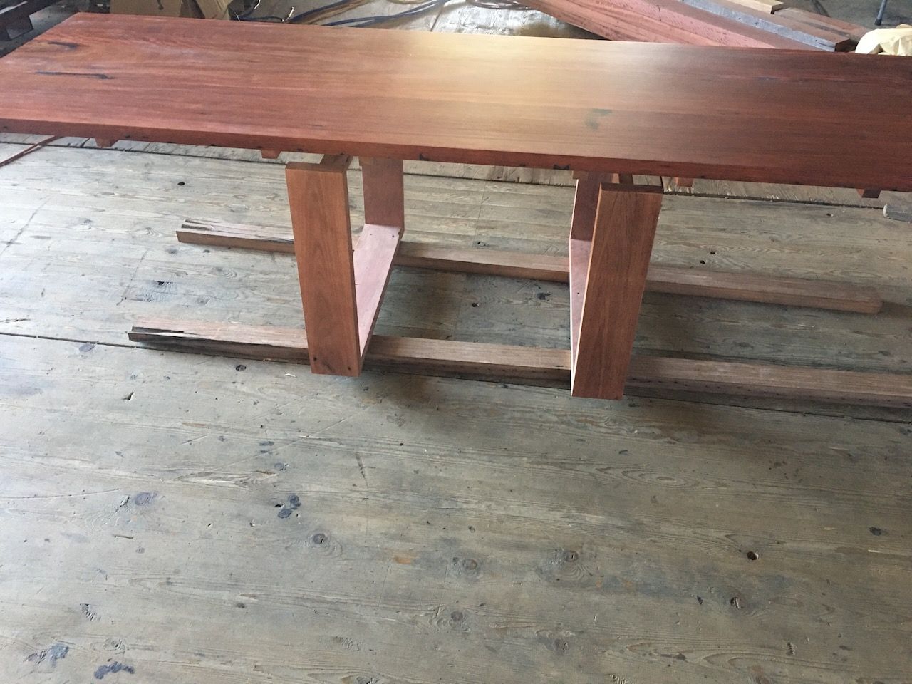 Wooden dining table with red-brown tabletop and supporting legs, on a concrete surface.