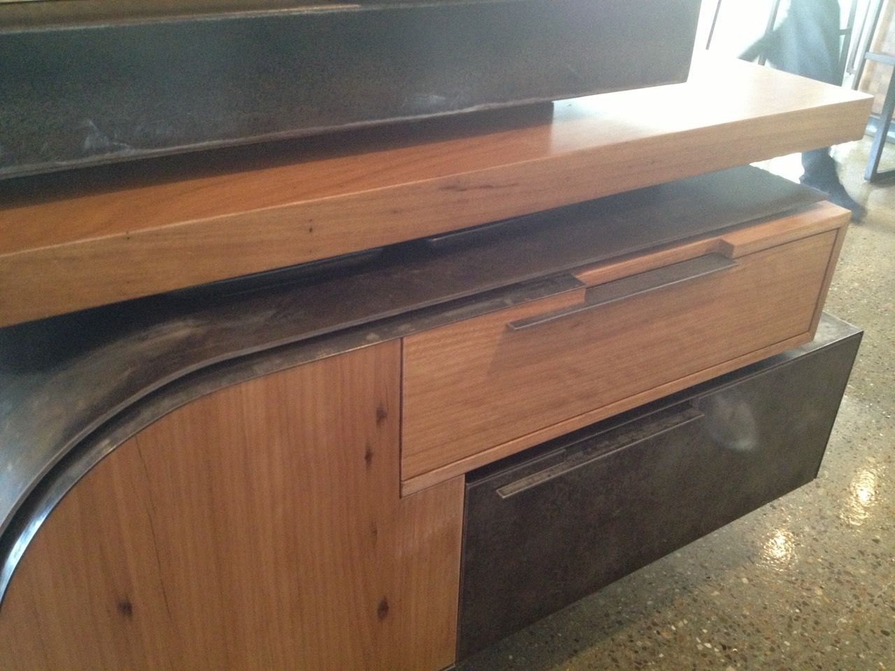Wooden cabinet with drawer, set on a dark, curved base. Shelf above with another dark surface.