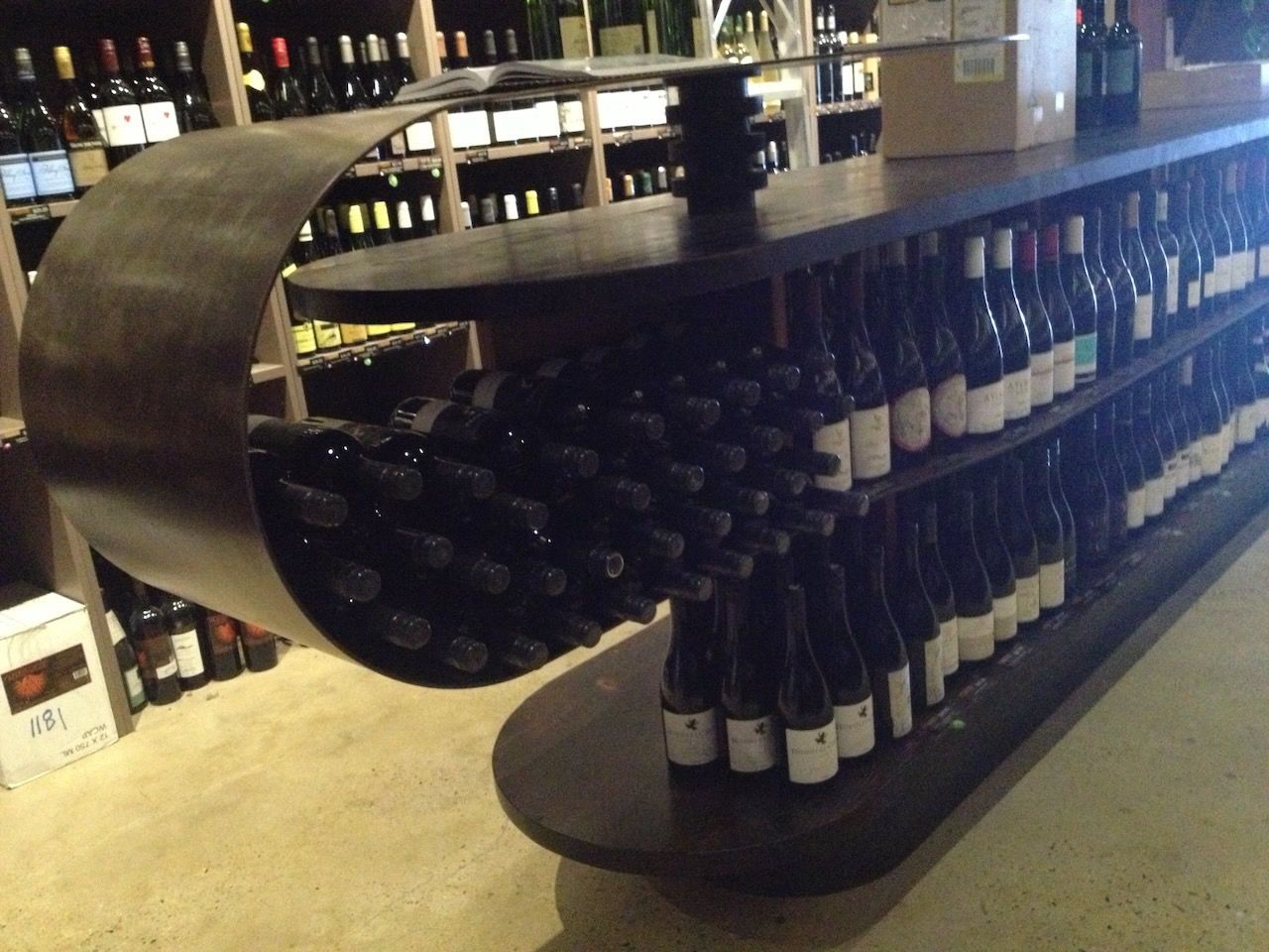 Dark wooden display with wine bottles. Curved shelf holds wine, with bottles also on lower shelves in wine shop.