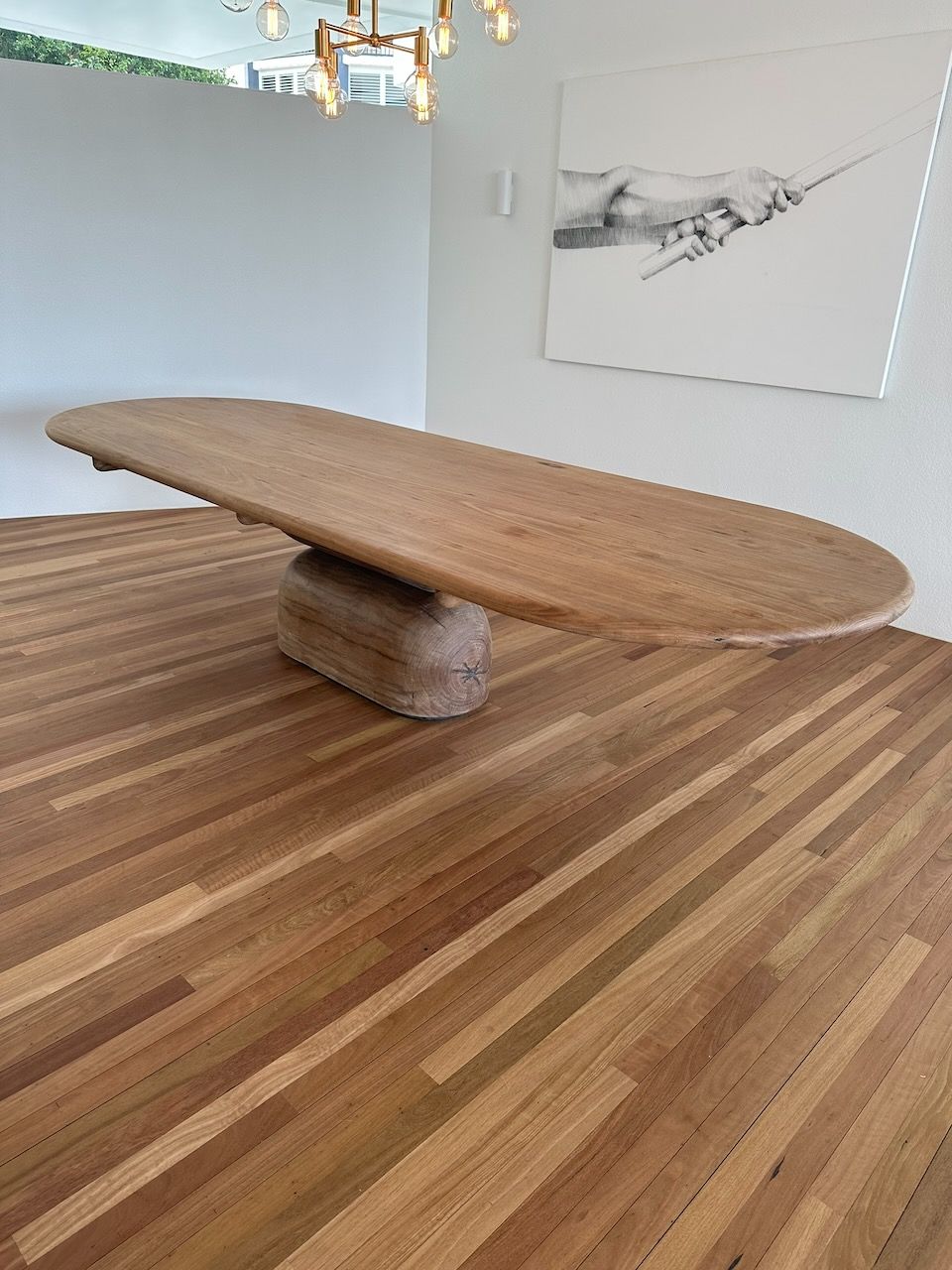 Oval wooden dining table on a cylindrical base, set on a hardwood floor. A painting hangs on the wall.