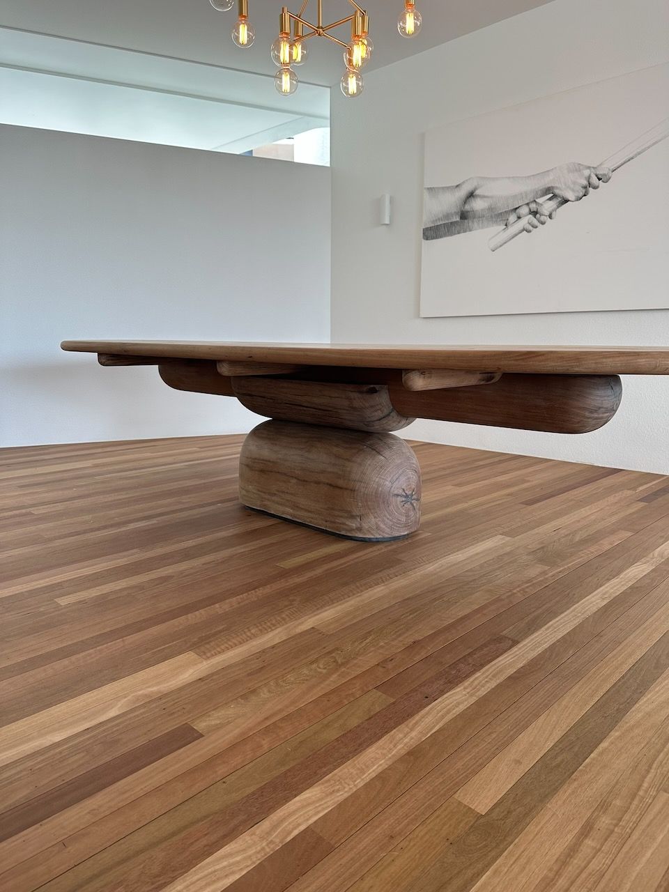 Wooden dining table with unique pedestal base on hardwood floor, with abstract artwork and chandelier.