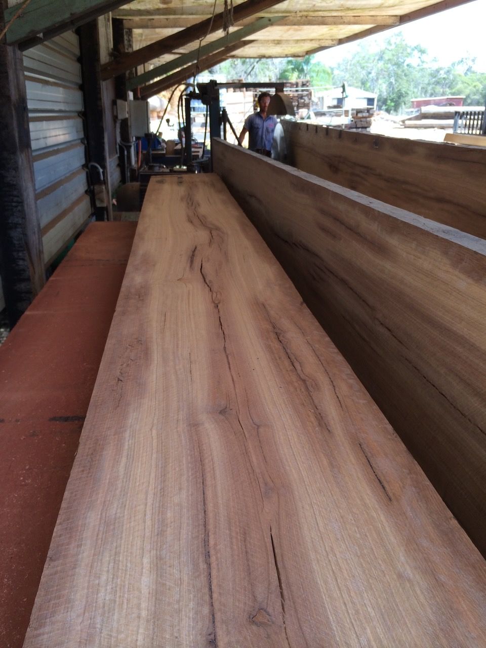 Large wooden planks in a sawmill, a person operating machinery in the background. Brown wood, outdoor setting.