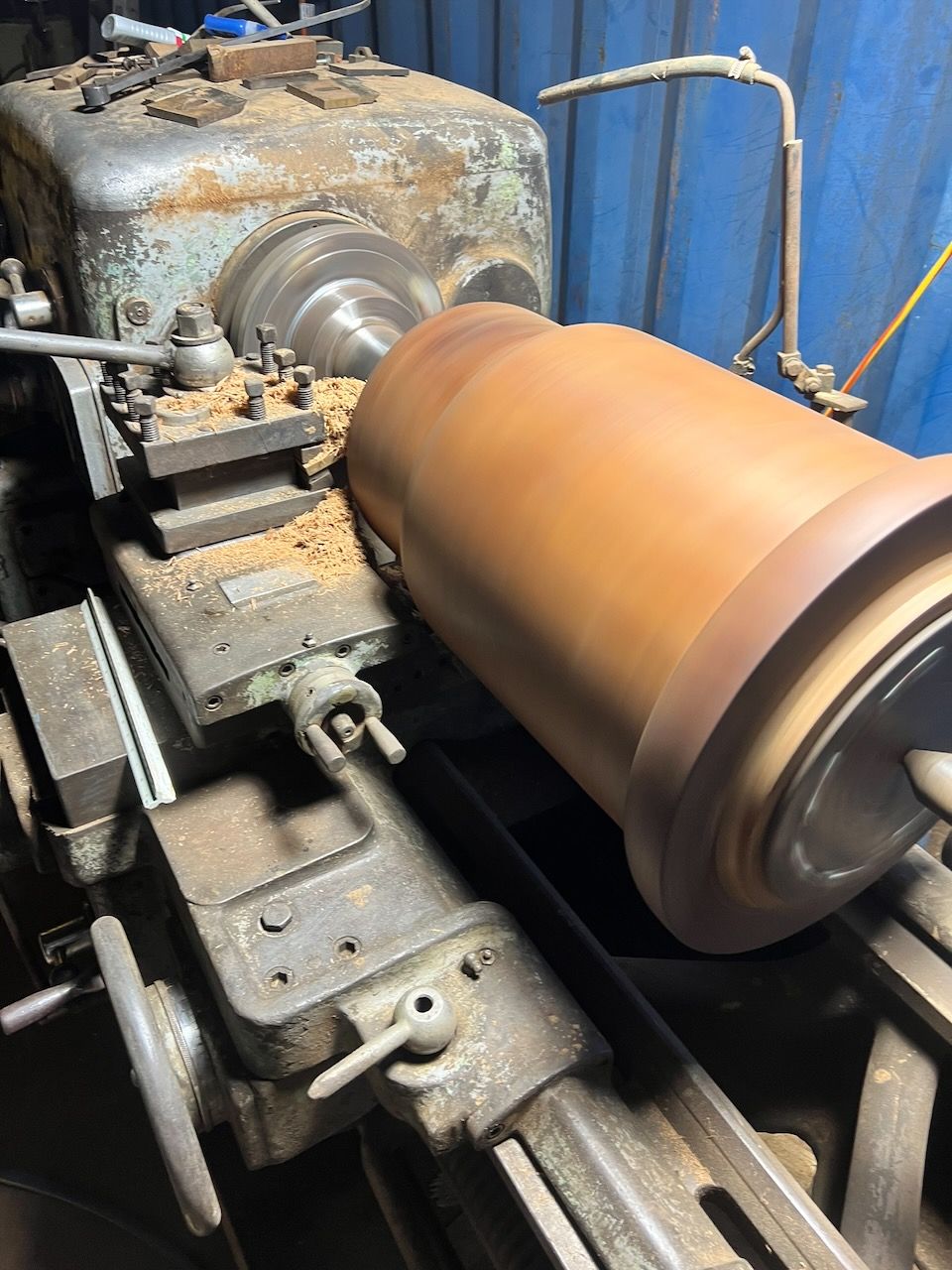A large wooden cylinder being turned on a lathe, shavings visible.