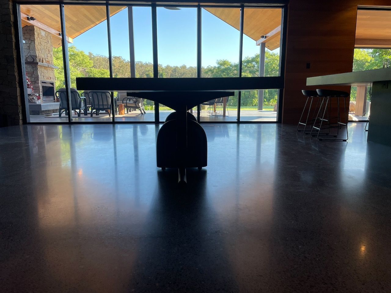 A modern dining table with a unique central pedestal base stands on a polished concrete floor before floor-to-ceiling windows.